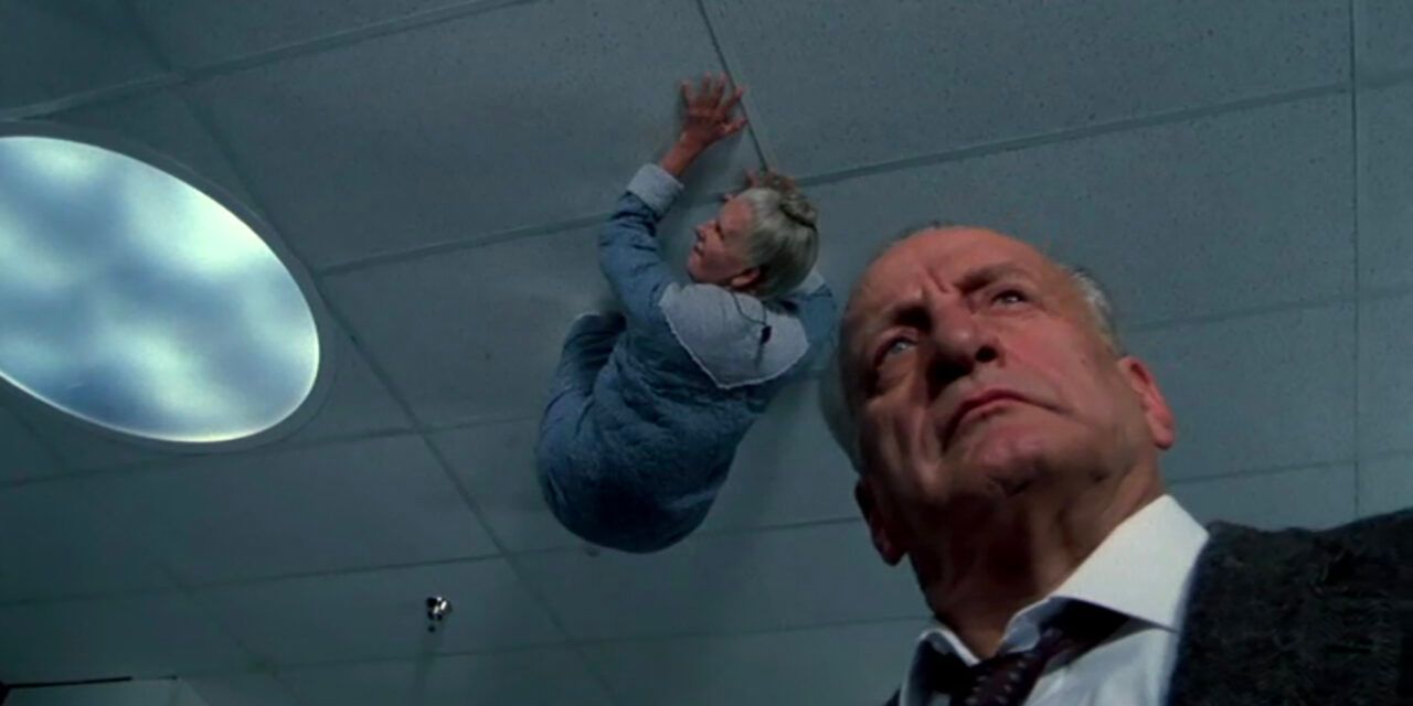 A man looks around a room while an old lady climbs on the ceiling above him.